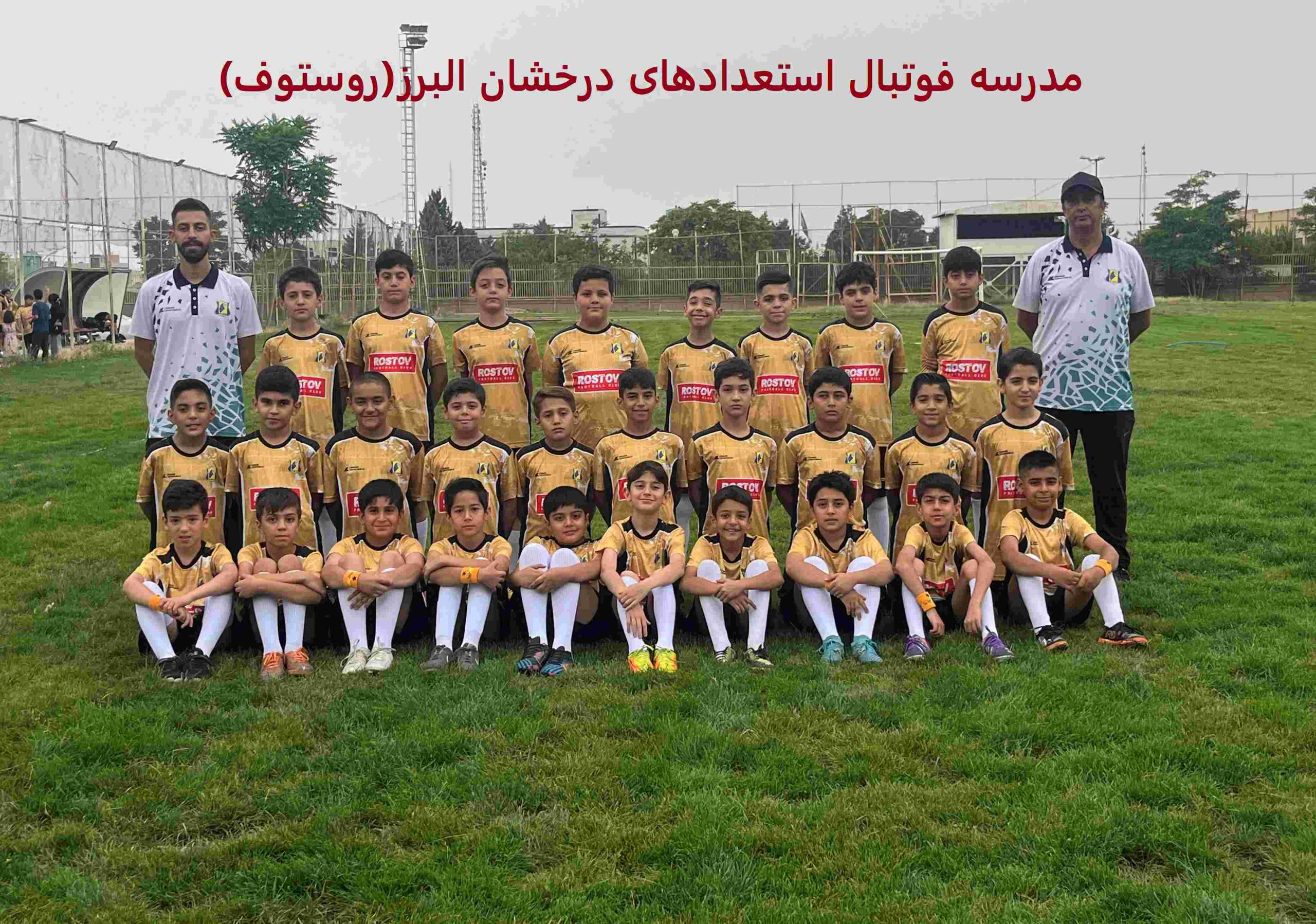 عکس گروهی از یک تیم فوتبال جوانان در کرج، شامل 16 بازیکن خردسال با لباس‌های زرد و مشکی با لوگوی "rostov"، ایستاده و نشسته روی چمن سبز زمین ورزشی، در کنار دو مربی با لباس‌های سفید و مشکی